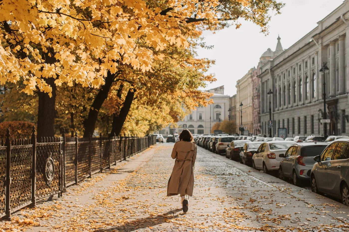 Woman in a trench coat walking along a leaf-covered sidewalk